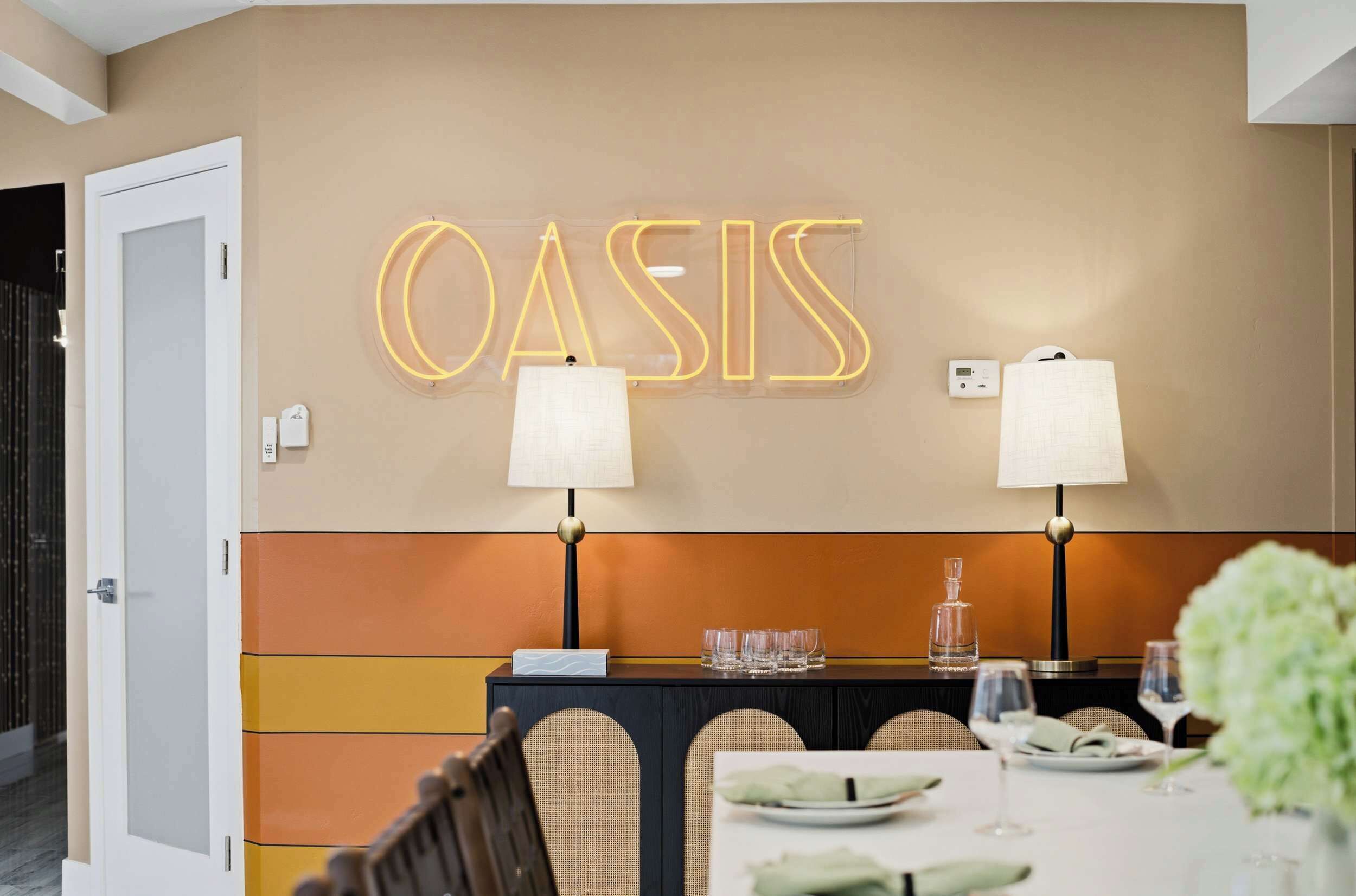 A hotel reception area with a neon sign reading "OASIS" above a reception desk. There are two lamps on either side of the desk, a white door to the left, and a dining area in the foreground with a table set for two.