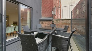 Eine Terrasse im Freien mit einem Glastisch und Korbstühlen, einer Ziegelwand und einem Metallzaun.
