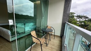 Een balkon met een glazen balustrade, rieten stoelen, een klein rond tafeltje en uitzicht op een zwembad en groen.