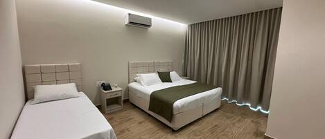 A hotel room with two single beds, a double bed, a nightstand with a lamp, and an air conditioning unit.