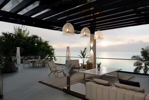 Een moderne buitenlounge met een glazen tafel, comfortabele zitplaatsen en uitzicht op de zee.