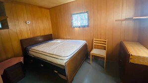 A small, single-bed room with wooden walls, a window with a patterned curtain, a wooden chair, and a small table with a lace tablecloth.