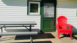 Una terraza de madera con una mesa de picnic y una silla roja frente a un edificio con puerta verde y revestimiento blanco.