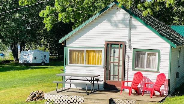 A small white cabin with a porch, a picnic table, and three red chairs.