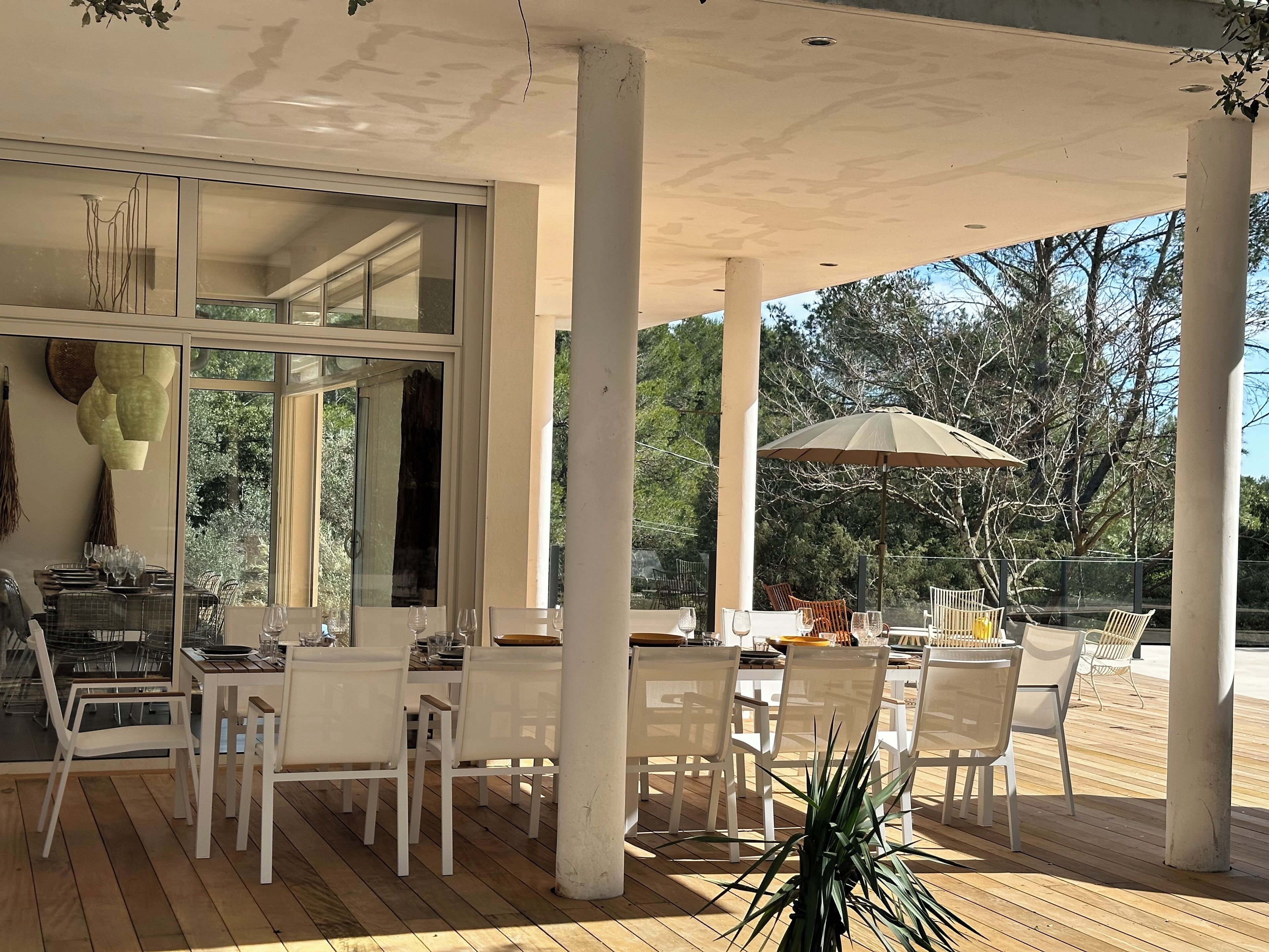 Un patio exterior con sillas y mesas blancas, un paraguas grande y vistas a árboles.