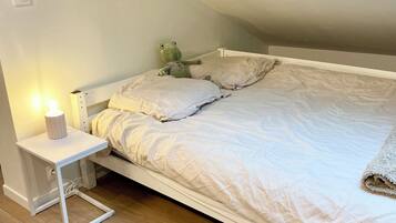 A small, neatly made bed with a white headboard and pillows, a bedside table with a lamp, and a wooden floor.