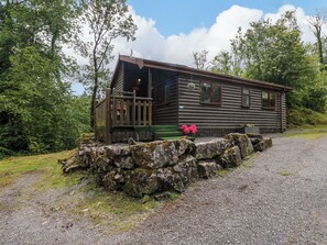 A log cabin with a porch, surrounded by a stone wall and greenery.
