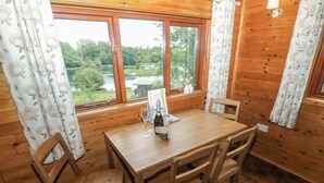 A wooden interior with a table and chairs, a window with a view of greenery, and a bottle on the table. - Ash Lodge (Llanelli)