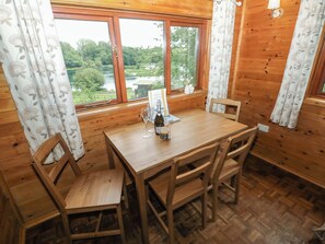 A wooden interior with a table and chairs, a window with a view of greenery, and a bottle on the table. - Ash Lodge (Llanelli)