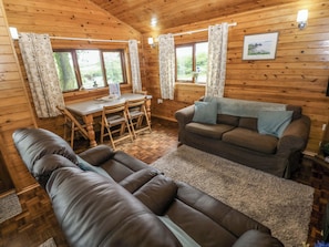 A wooden cabin interior with a sofa, a dining table with chairs, and a window with curtains.