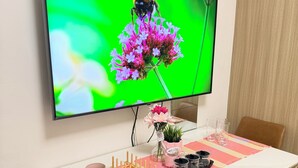 Un coin repas avec une table dressée pour deux personnes, un téléviseur fixé au mur qui diffuse une image de fleurs, et une horloge au-dessus.