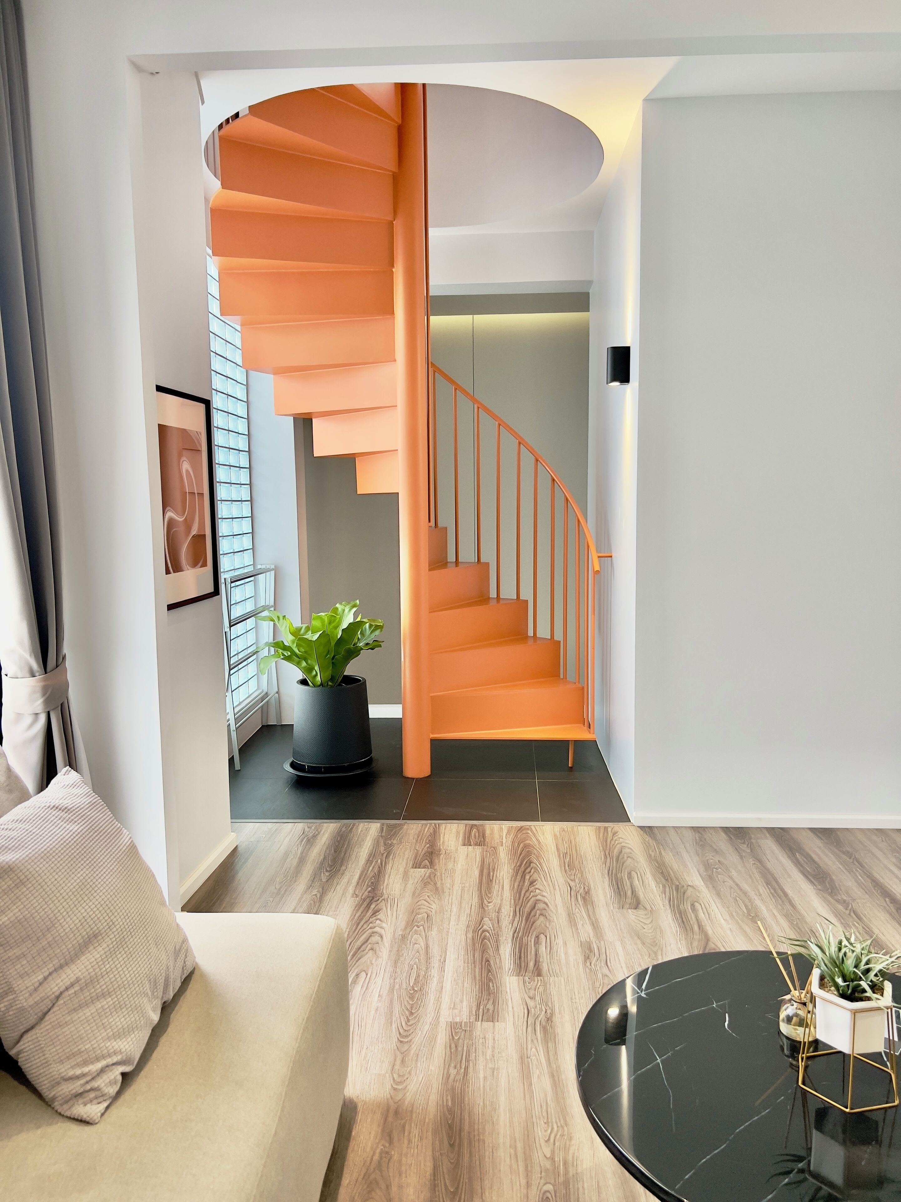 A modern living room with a wooden floor, a round black coffee table, a beige sofa, and a spiral staircase with orange railings.