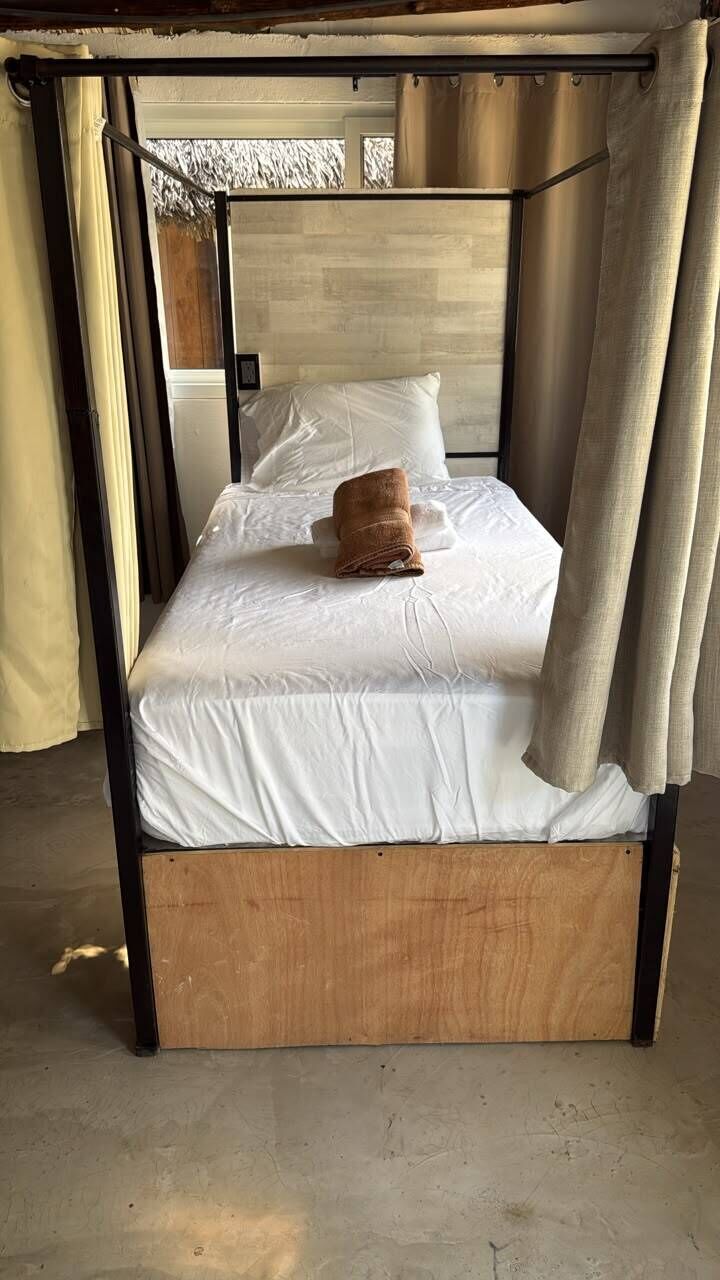 Una cama individual con estructura y cabecero de madera, una toalla marrón sobre la cama y cortinas color beige.