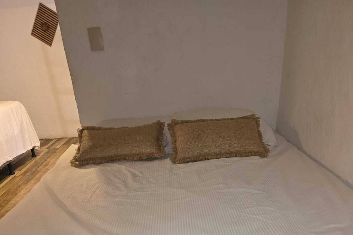 Una cama con ropa blanca y dos almohadas color beige en una habitación con piso de madera y paredes blancas.