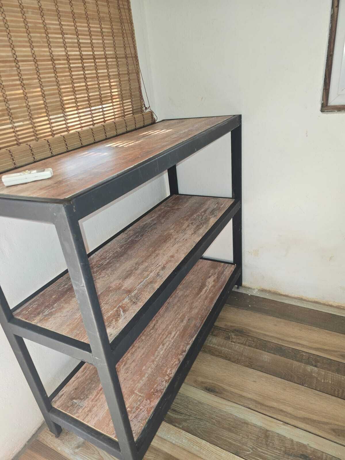 A metal-framed shelf with wooden slats, positioned against a wall with a window with blinds.