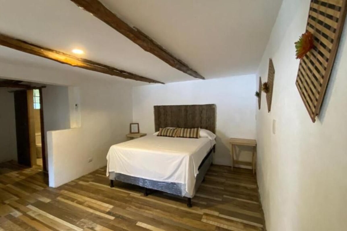 A bedroom with a wooden floor, a bed with a white bedspread, a wooden headboard, a nightstand, and a wall-mounted shelf with decorative items.