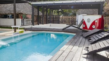 A swimming pool with a wooden deck, a thatched-roof structure, and a VW camper.
