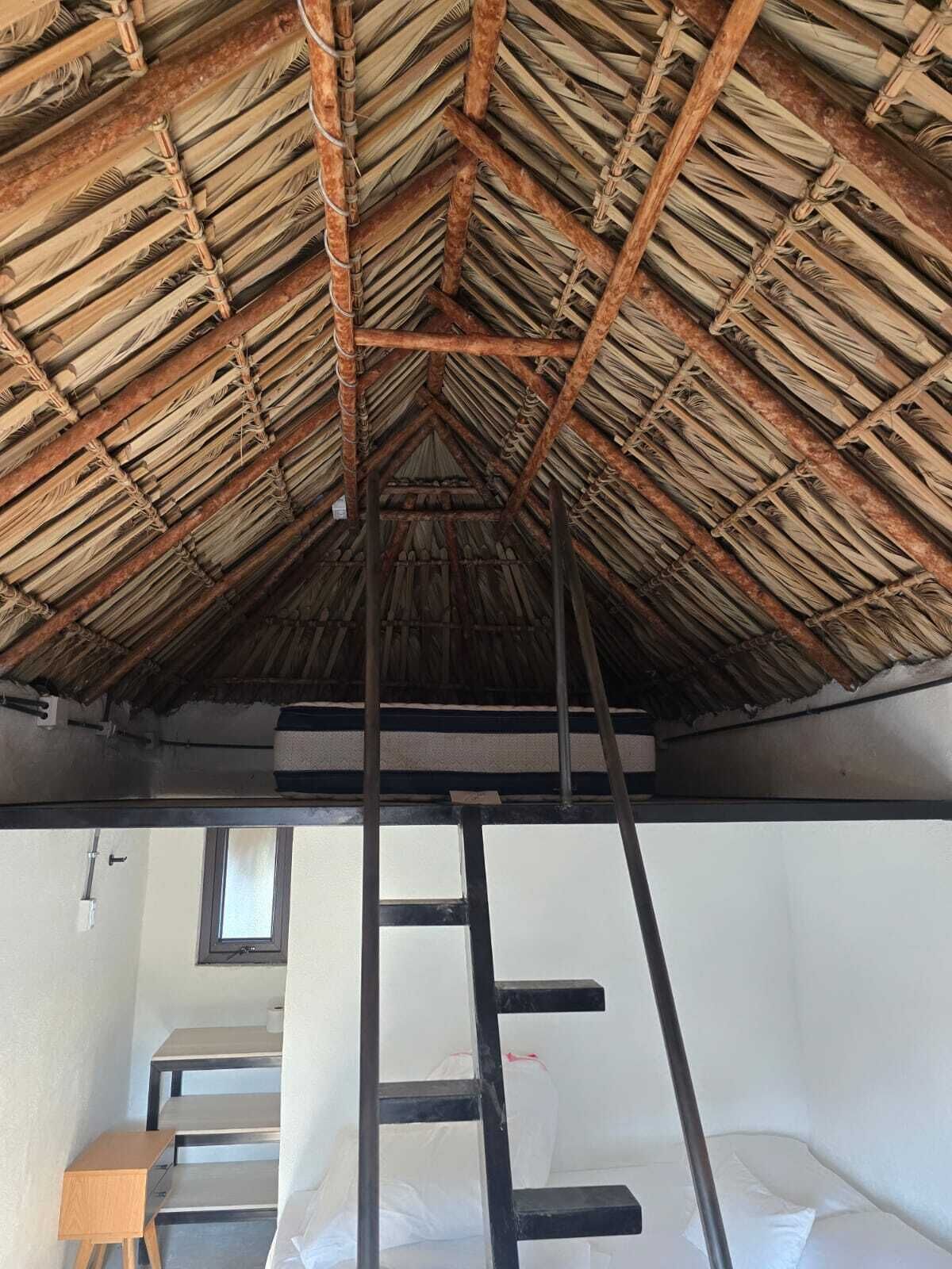 Una habitación con techo de paja, literas, una escalera y un ventanal.