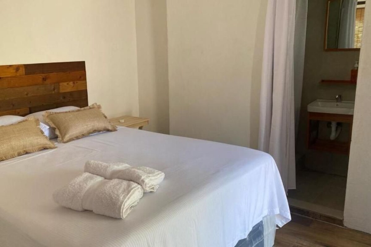 Una cama bien hecha con sábanas blancas y dos almohadas beige, un cabecero de madera y una mesita de noche con una lámpara.