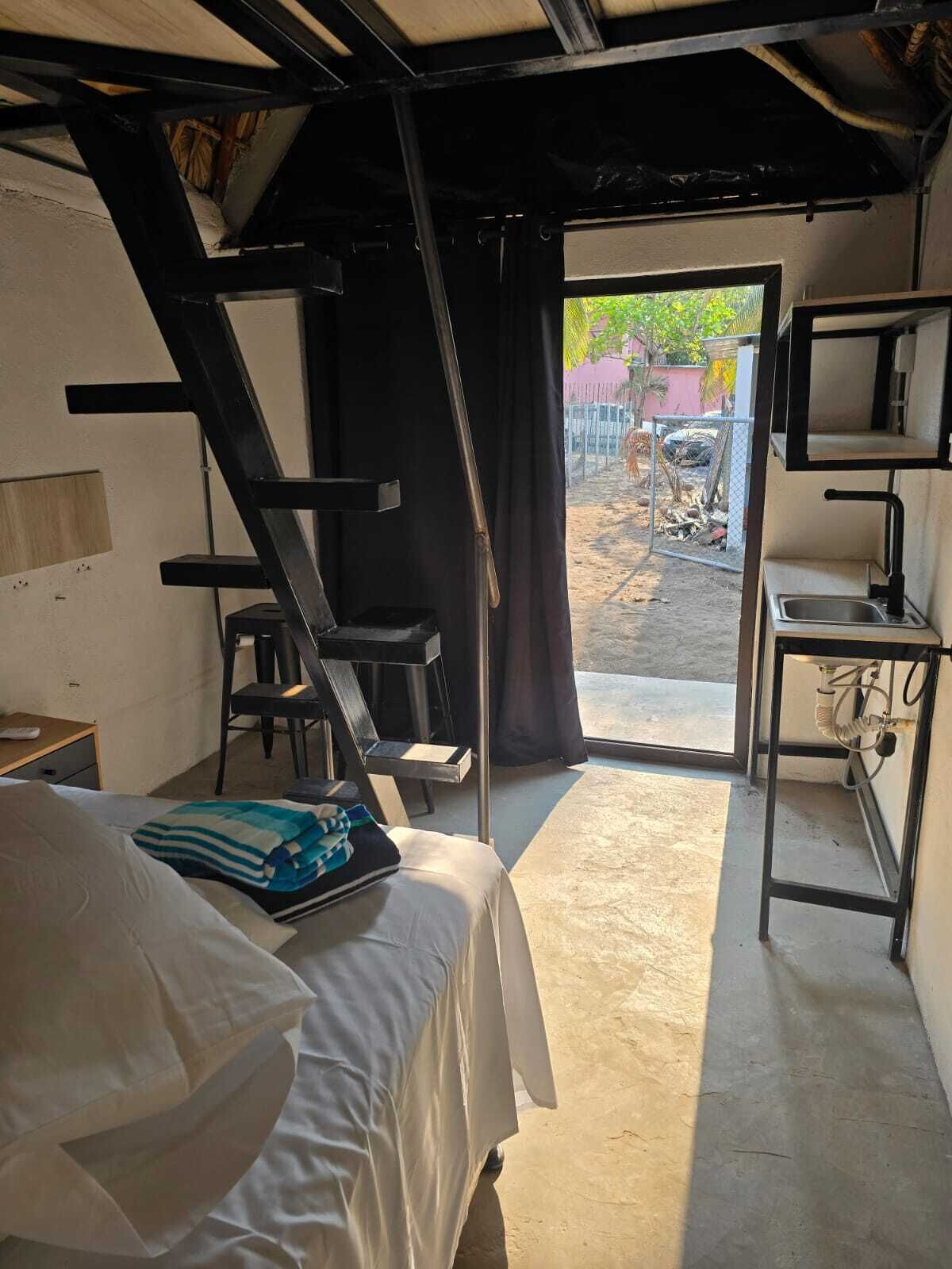 Ein kleines Zimmer mit Bett, Schreibtisch, Stuhl, Fenster und einer Tür, die nach draußen führt.