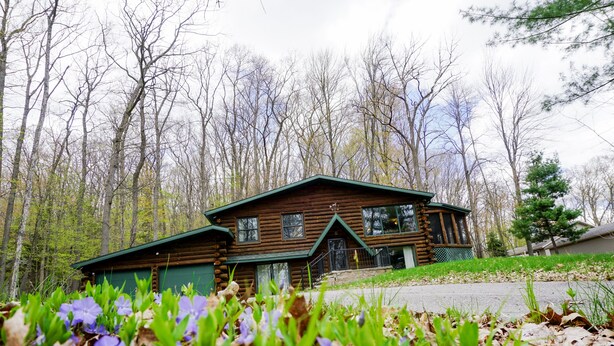 A log cabin with a green roof, surrounded by trees and purple flowers. - Rib Mountain Cabin (Wausau)
