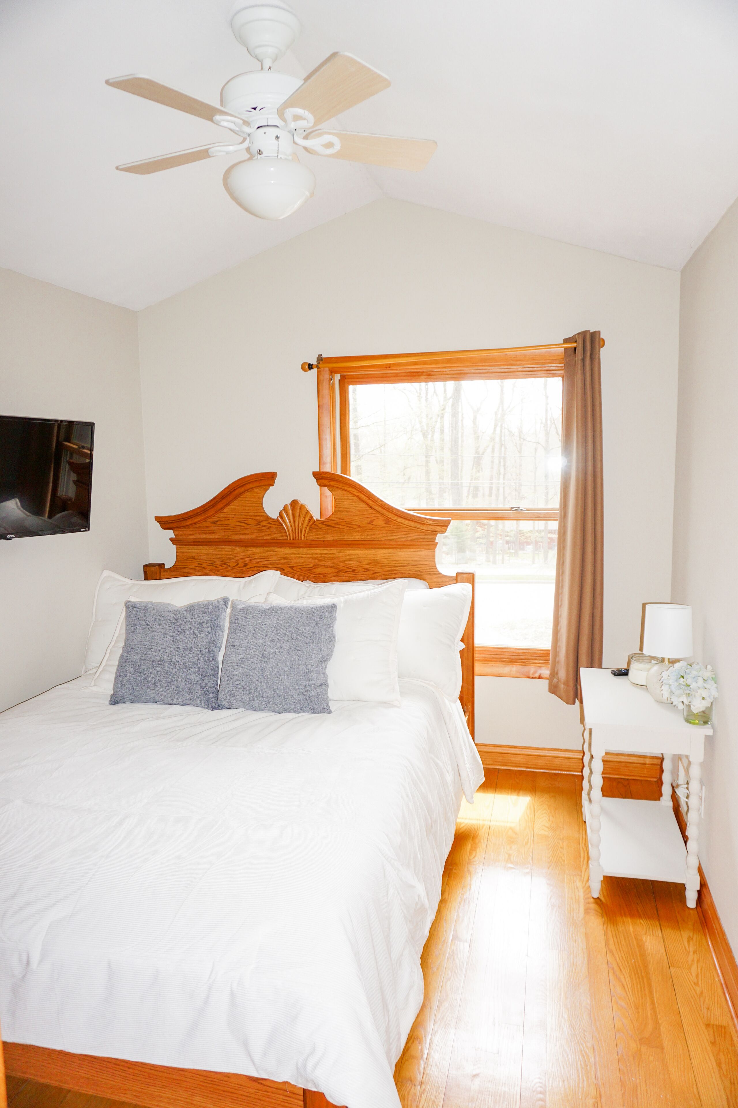 A bedroom with a wooden bed, a ceiling fan, a window with curtains, a bedside table, and a TV mounted on the wall.
