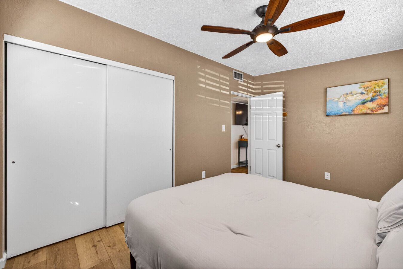A bedroom with a bed, a ceiling fan, a white door, and a painting on the wall.