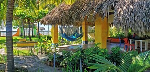 Un espacio de descanso al aire libre con techo, una hamaca, palmeras y un edificio amarillo.