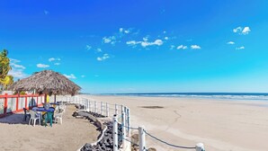 En strand med en struktur med stråtækte tage, hvide stole og et rødt hegn.
