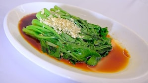 Ein weißer, ovaler Teller mit einer Portion grüner Blattgemüse, möglicherweise Bok Choy, belegt mit gehacktem Knoblauch und einer dunklen Soße.