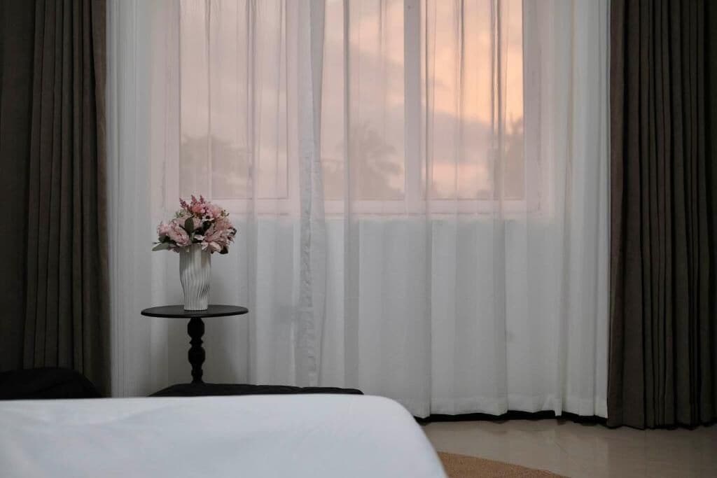 Una camera d'albergo con un letto, un tavolino rotondo con un vaso di fiori e tende leggere.