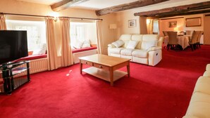 A cozy living room with a red carpet, a white sofa, a wooden coffee table, a TV, and a dining area with a table and chairs. - The Farmhouse (Keswick)