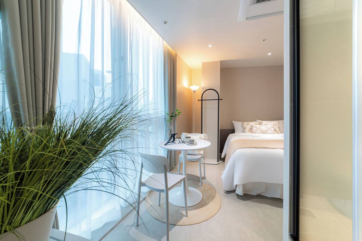 Una habitación de hotel moderna con cama, mesita de noche, una silla y un ventanal con cortinas translúcidas.