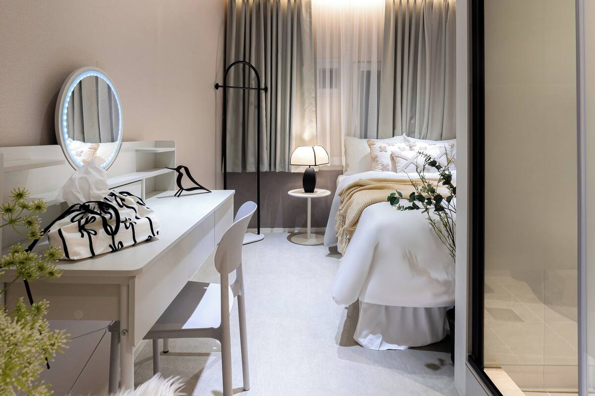 Una habitación de hotel moderna con un escritorio blanco, un espejo, una cama con ropa de cama blanca, una mesita redonda pequeña con una lámpara y un ventanal grande con cortinas.