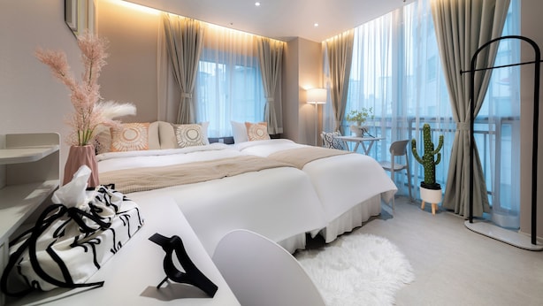Ein modernes Schlafzimmer mit einem großen Bett, einem Nachttisch, einer Lampe, einer Zimmerpflanze und Blick auf die Stadt durch große Fenster.