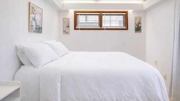Ein ordentlich bezogenes Bett in einem Zimmer mit weißen Wänden, einem Fenster mit Jalousien und zwei gerahmten Kunstwerken an der Wand.