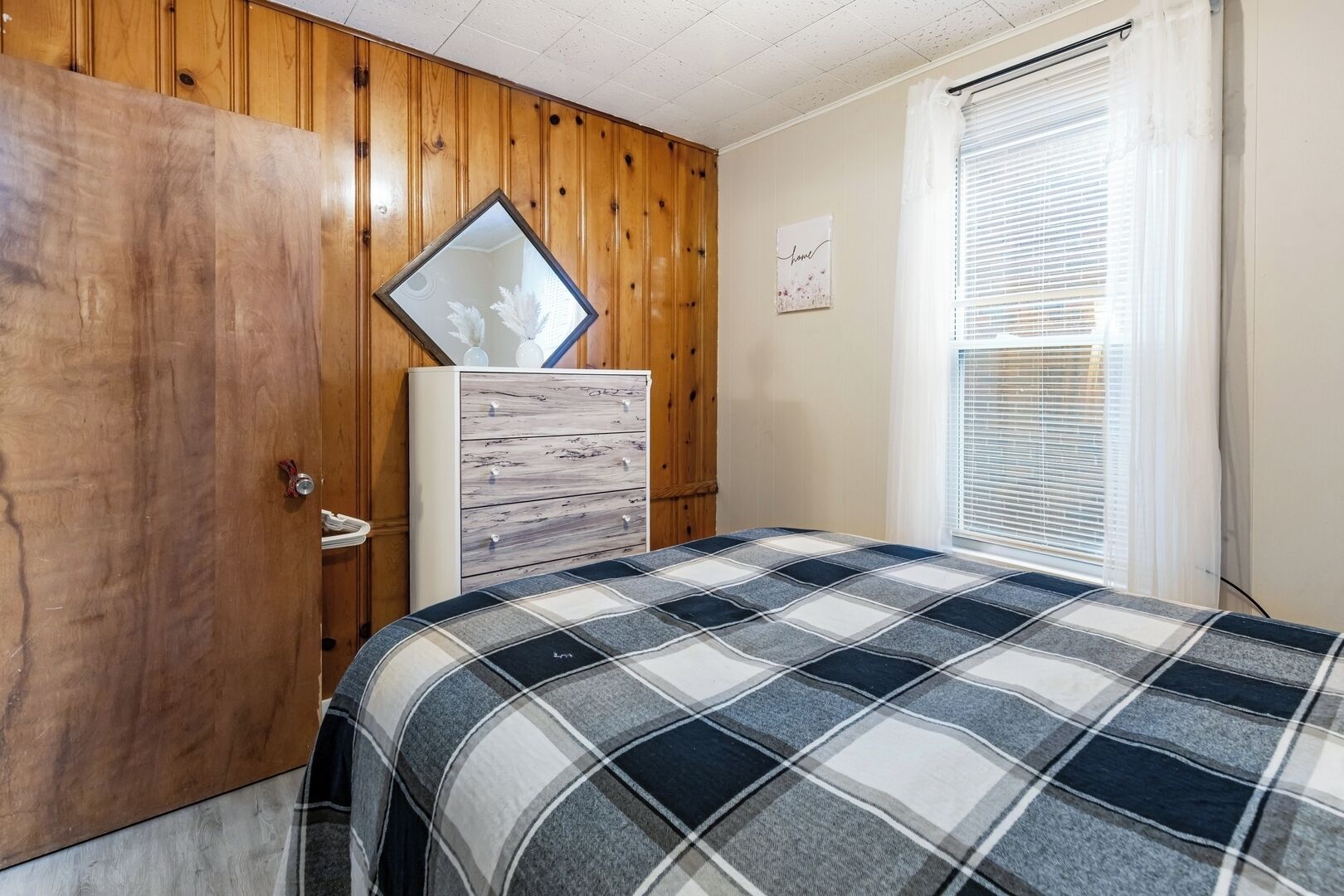 Une chambre avec une armoire en bois, une commode, un lit recouvert d’une couverture à carreaux, une fenêtre avec des stores et un mur orné d’un panneau indiquant « chez soi ».