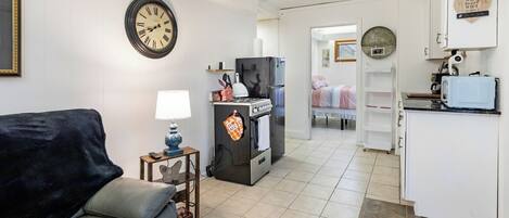 A compact living space with a sofa, a small table with a lamp, a kitchen area with a refrigerator, oven, and microwave, and a clock on the wall.
