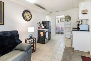 A compact living space with a sofa, a small table with a lamp, a kitchen area with a refrigerator, oven, and microwave, and a clock on the wall.