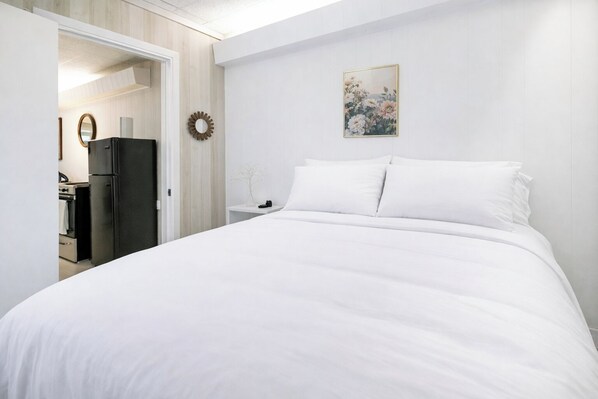 A hotel room with a large bed, a small bedside table, a wall-mounted artwork, and a view into the kitchen area.