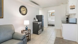 Ein kompakter Wohnbereich mit Sofa, einem kleinen Tisch mit Lampe, einer Küchenzeile mit Kühlschrank, Herd und Mikrowelle sowie einer Uhr an der Wand.