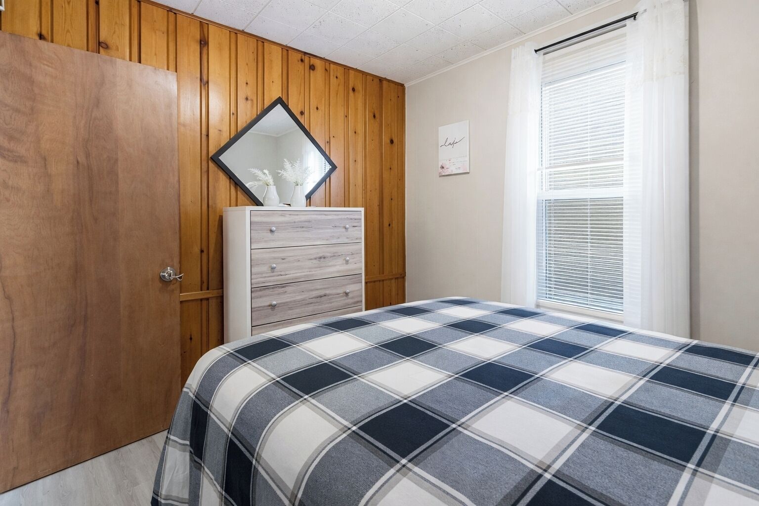 Ein Schlafzimmer mit einem Holzschrank, einer weißen Kommode, einem Bett mit kariertem Überwurf, einem Spiegel und einem Fenster mit Jalousien.