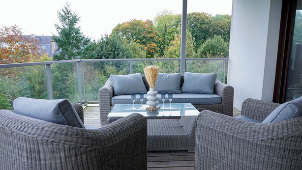 A balcony with wicker furniture, a glass table, and a vase with dried plants.