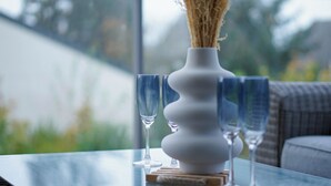 A clear glass table with a white vase containing dried plants, two empty wine glasses, and a wooden coaster.