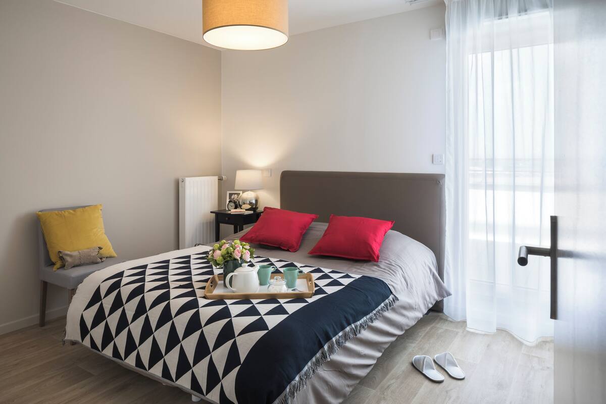 A modern hotel room with a large bed, a bedside table, a nightstand, a window with sheer curtains, and a pair of slippers on the floor.