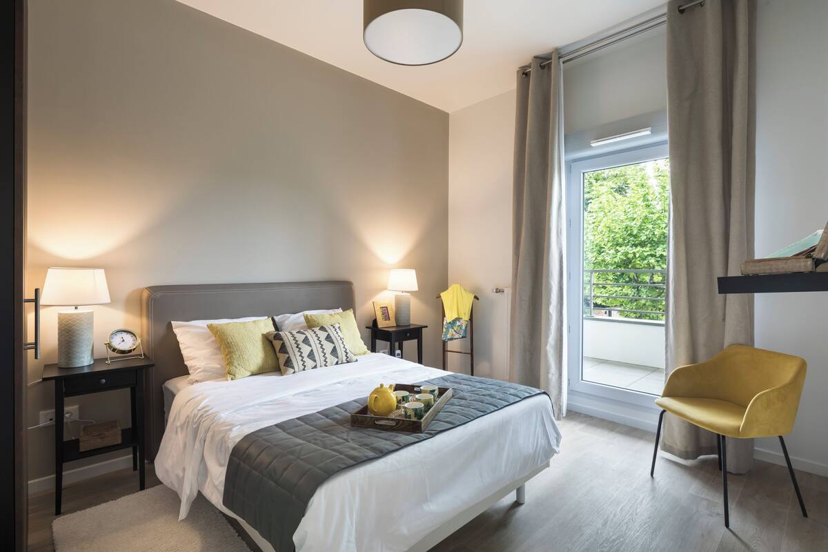 A modern hotel room with a large bed, bedside lamps, a small table, a chair, and a window with curtains.