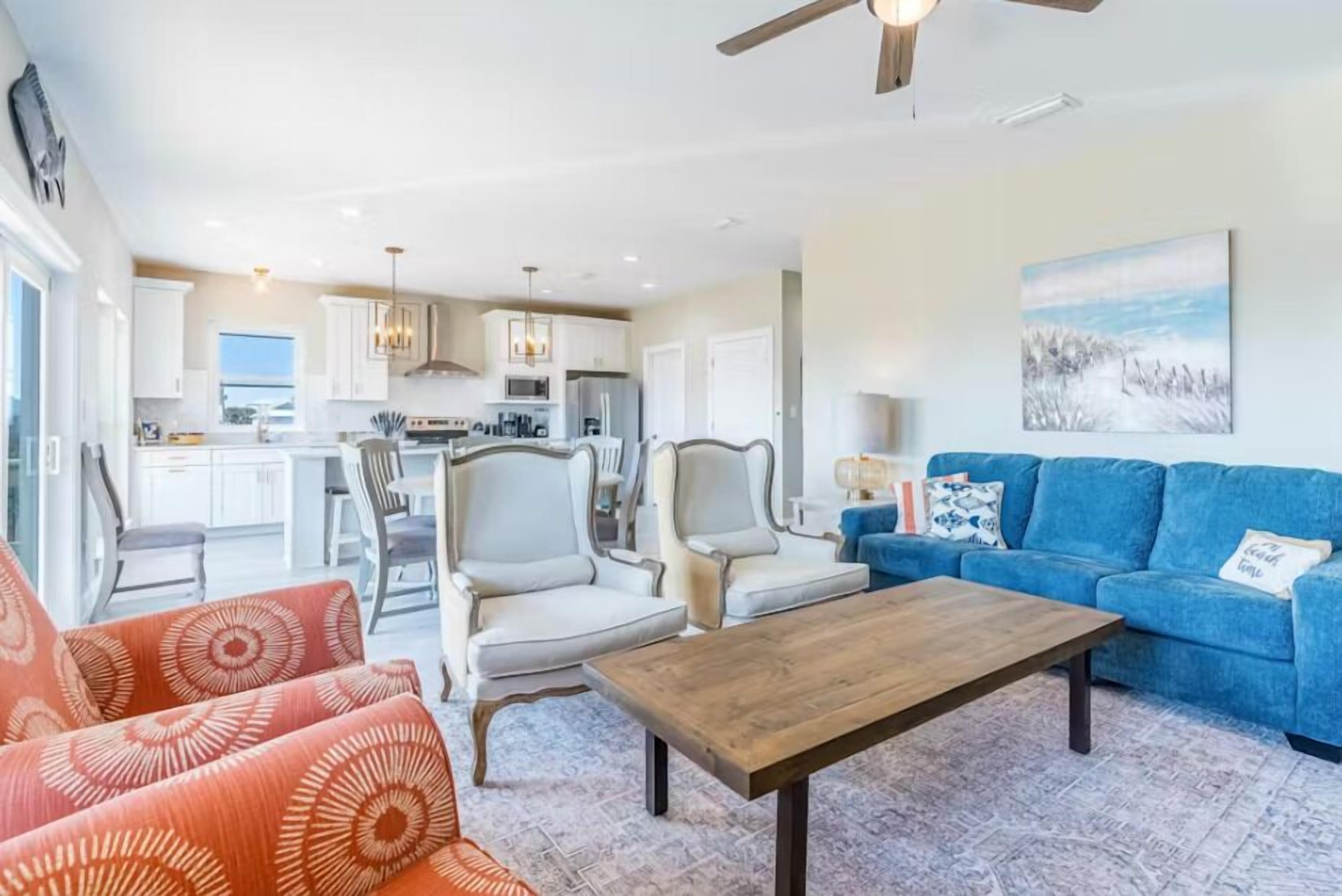 A spacious living room with a blue sofa, orange patterned sofa, and a wooden coffee table. There is a dining area with chairs and a kitchen in the background.