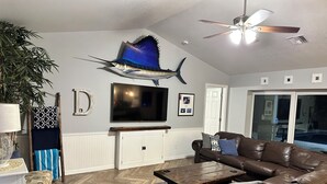 Ein Wohnzimmer mit braunem Ledersofa, einem Couchtisch, einem an der Wand montierten Fernseher, einem Deckenventilator und einer großen Fischdekoration.