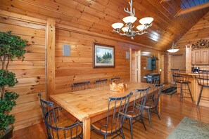 A wooden cabin interior with a large dining table, chairs, a chandelier, framed picture, and a kitchen area. - Spectacular 4BR Cabin w/ Game Room, Hot Tub, Views (Gatlinburg)