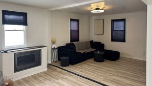 A modern living room with a black sectional sofa, a small side table, and a radiator. There are three windows with blinds, a ceiling light fixture, and a framed artwork on the wall. - Beautiful 2-bedroom house in tranquil Warren perfect for relaxing stays (Warren)